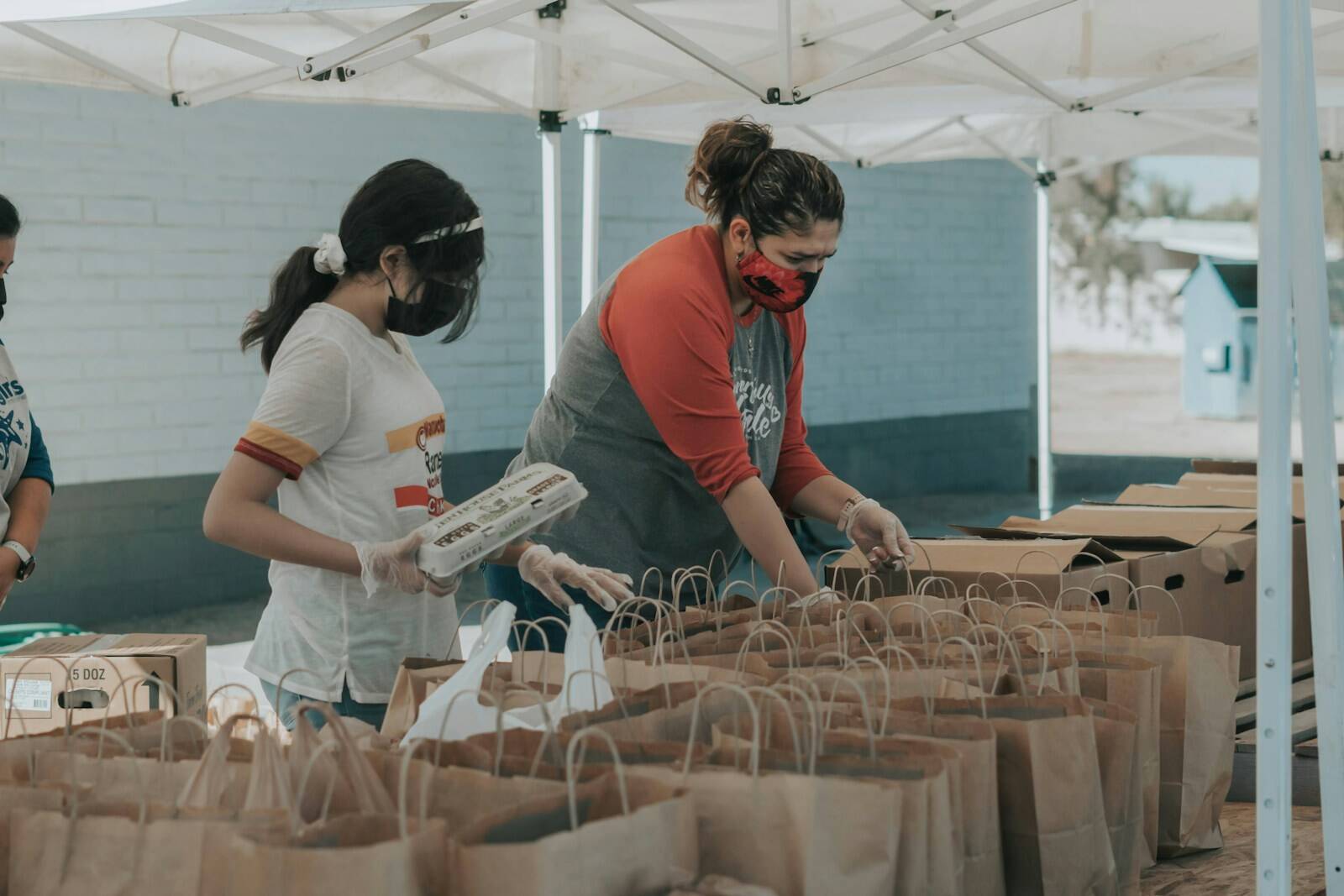 Two women wearing face masks and gloves packing paper bags under a white canopy
